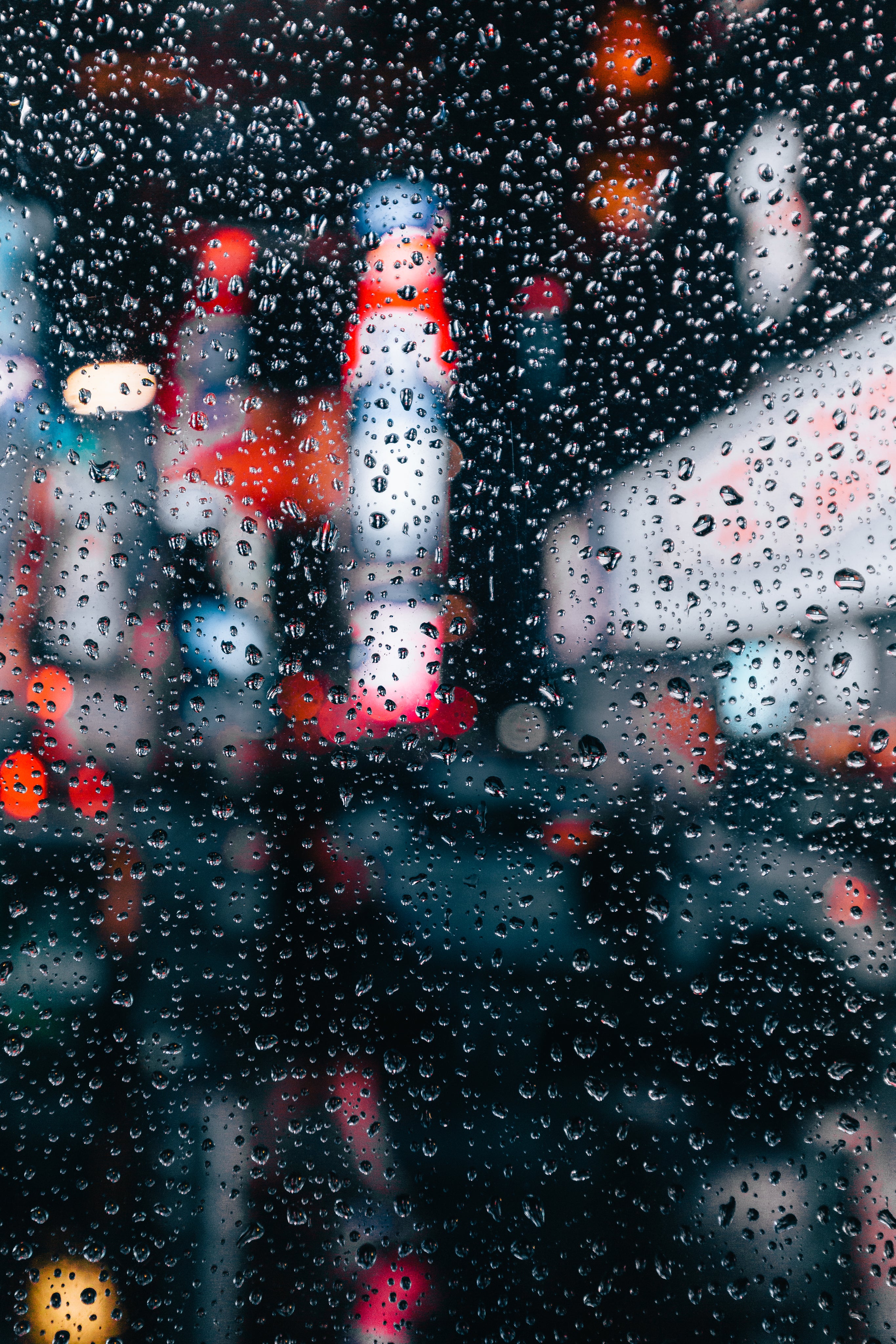 Raindrops cover a window, blurring the view of bright city lights and colorful signs outside, creating a vibrant and moody urban scene.
